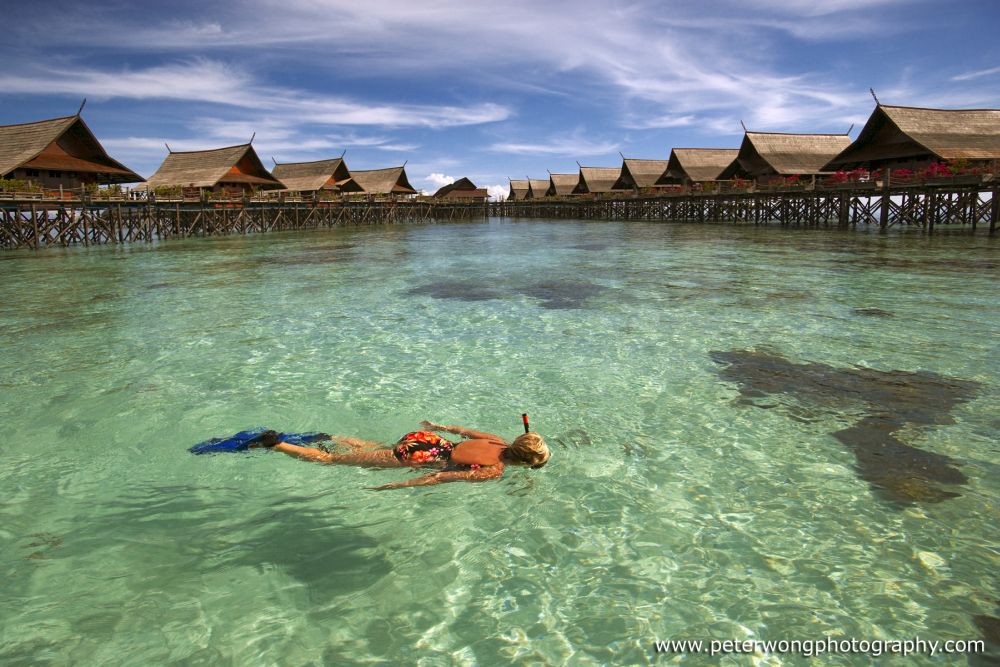 Snorkelers swimming over a shallow, healthy coral reef in the stunningly clear waters in front of the Kapalai resort chalets.