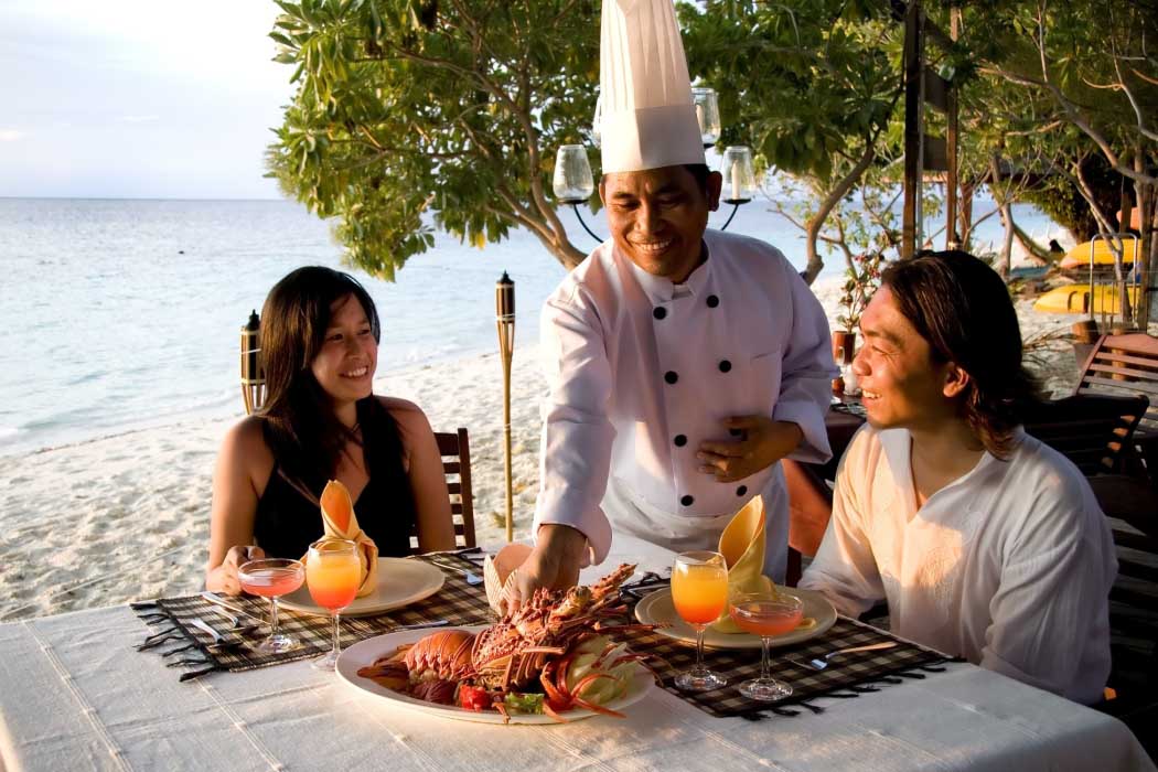 Couple enjoying a private romantic dinner on the beach at Mataking Island Resort, as a chef presents them with a fresh lobster dish at sunset.
