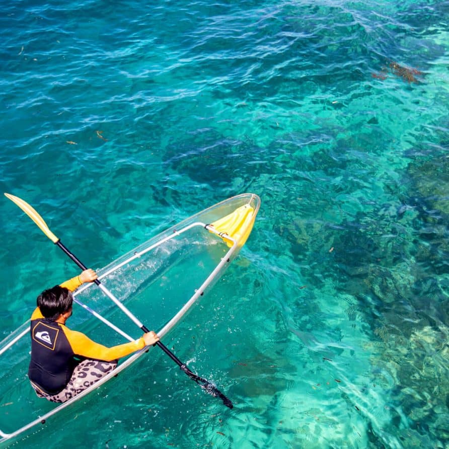 Guest relaxing in a transparent kayak, looking down at the beautiful coral reef below in the crystal-clear waters of Mabul island.